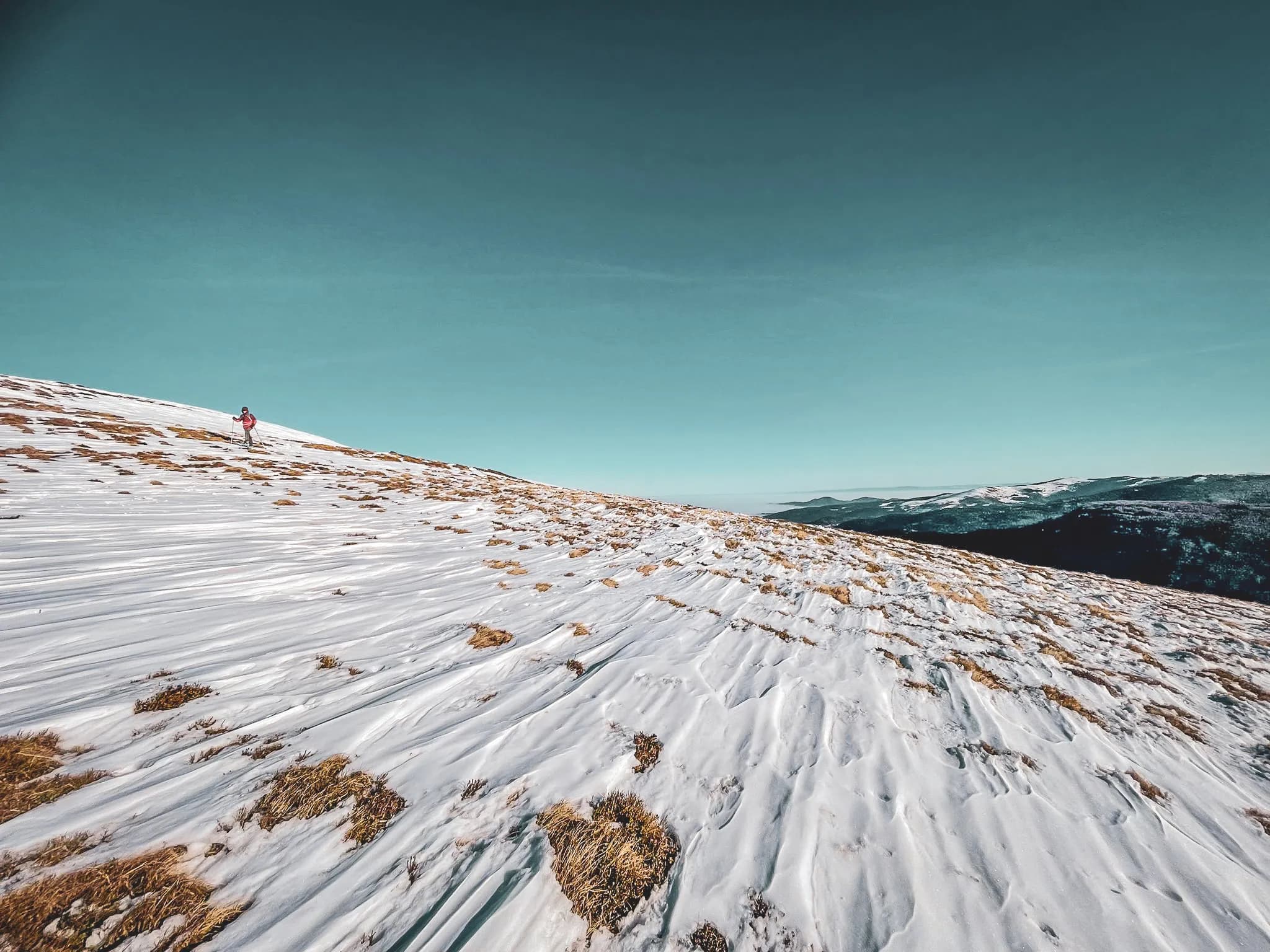 Een berglandschap waarin een persoon met een rode jas over een besneeuwde helling loopt. De golvingen van de sneeuw creëren interessante patronen, terwijl hier en daar vegetatie tevoorschijn komt.