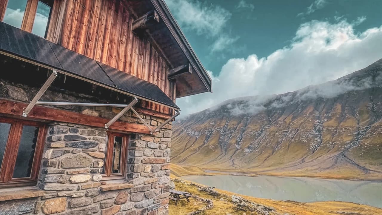 Un chalet en bois face à des sommets majestueux, accompagnés d'un lac serein et de nuages.