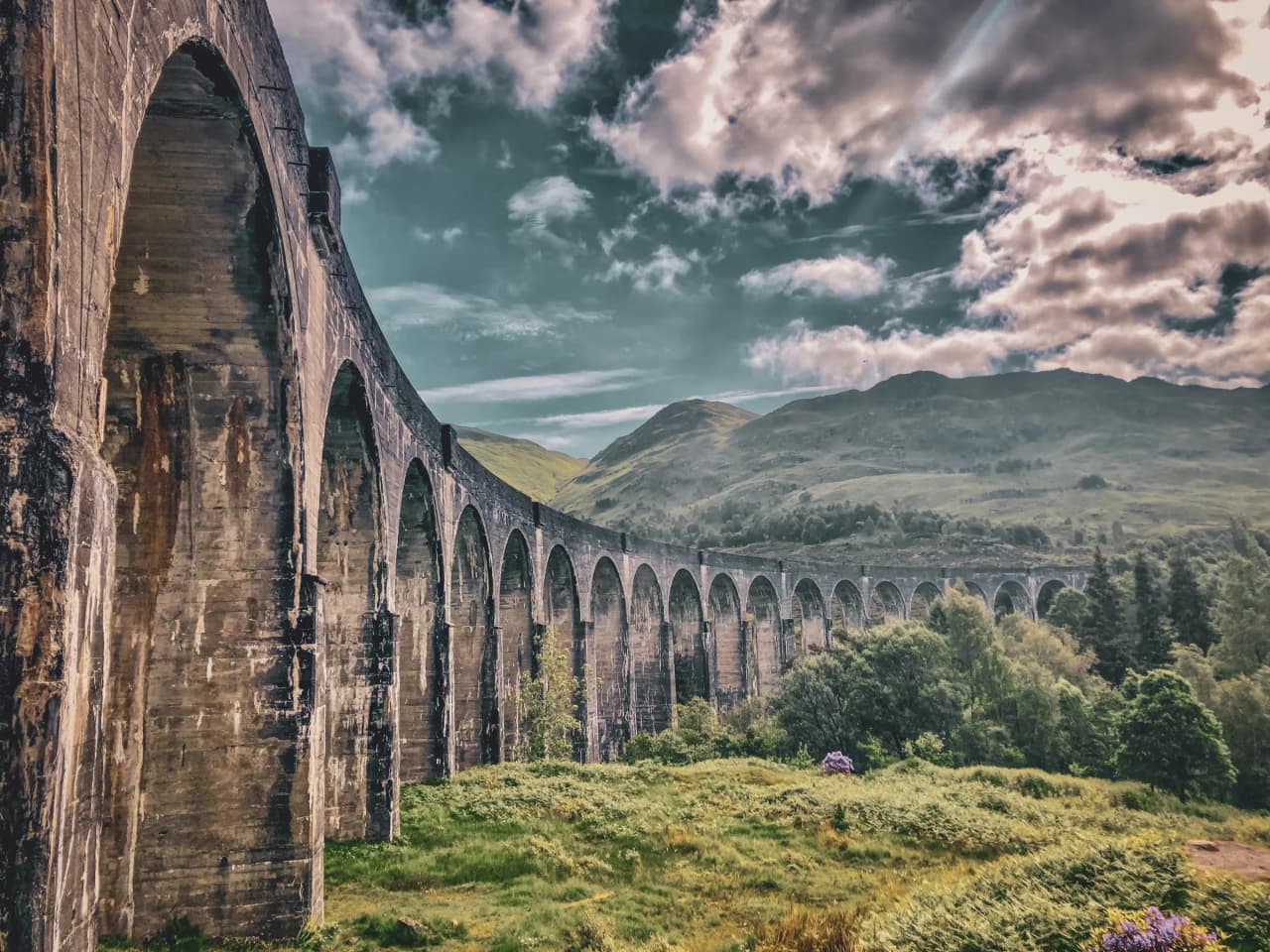 A majestic viaduct lined with greenery, set against a dramatic Scottish Highlands sky.