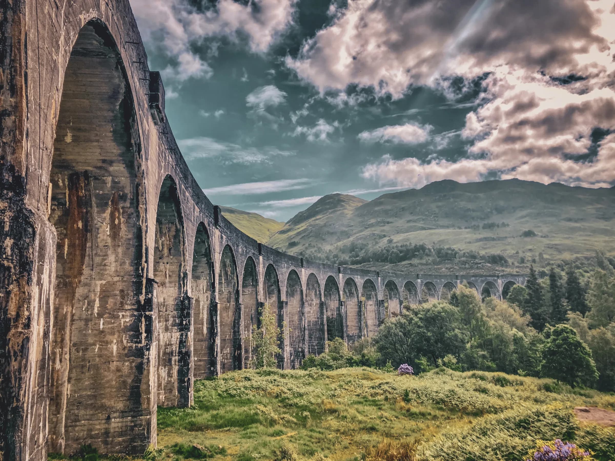 A majestic viaduct lined with greenery, set against a dramatic Scottish Highlands sky.