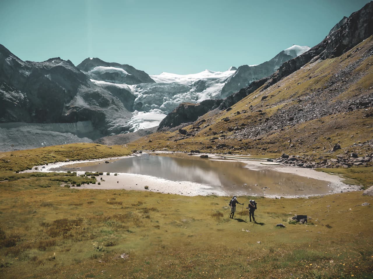 Two hikers stand near an Alpine lake, with the Aletsch glacier in the background.