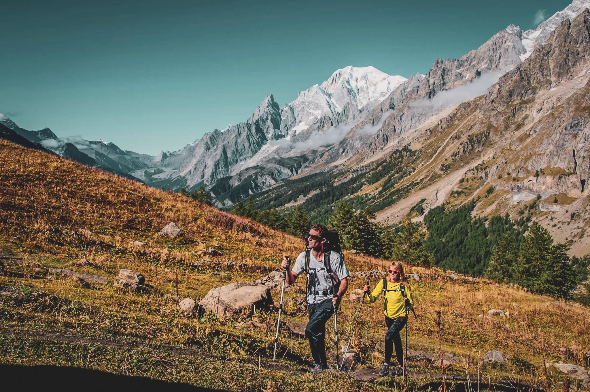 Un paysage montagneux avec des pics enneigés en arrière-plan, deux randonneurs avançant sur un chemin dans une vallée verdoyante. Le randonneur adulte porte un sac à dos et des lunettes de soleil, tandis qu'un