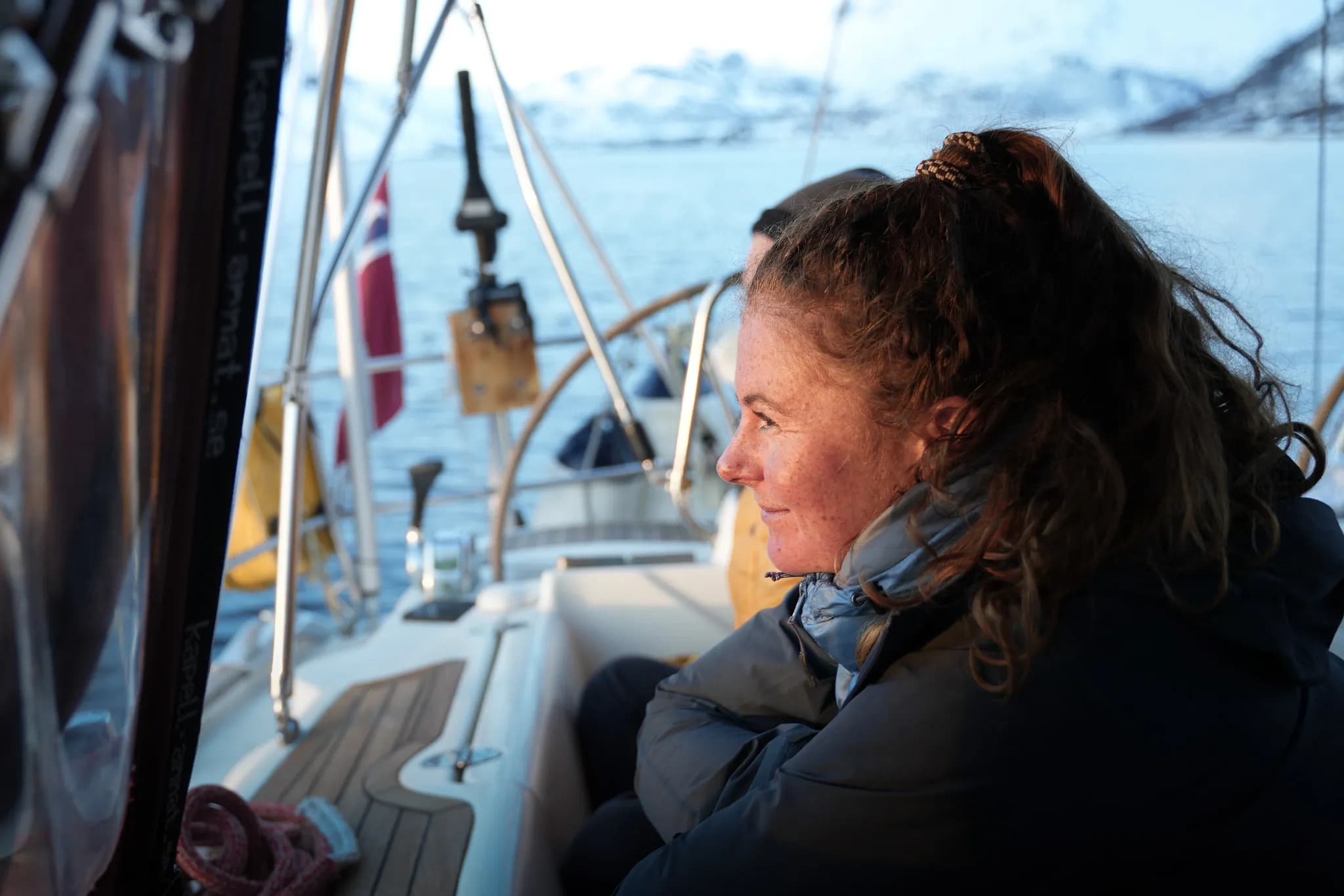 Un bateau de plaisance navigue sur une mer calme, sous un ciel clair. Une femme, aux cheveux frisés et attachés, est assise sur le bord, observant l'horizon. Elle porte une veste épaisse, suggérant