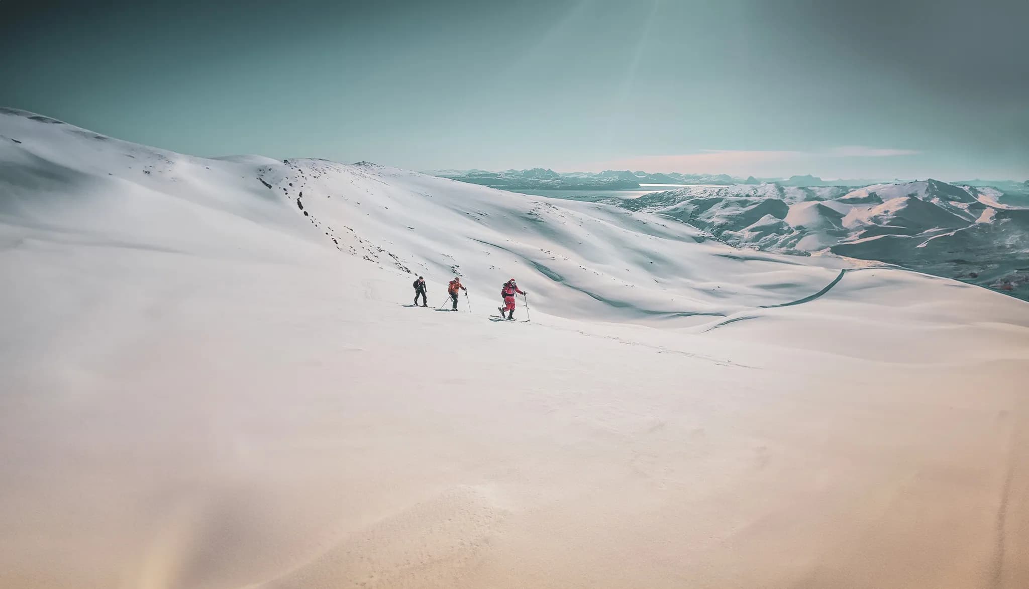 Skiërs op een uitgestrekte vlakte van ongerepte sneeuw, omringd door glooiende heuvels en bergen op de achtergrond. De lucht is helderblauw met zacht licht, wat een serene sfeer creëert. Bezoek