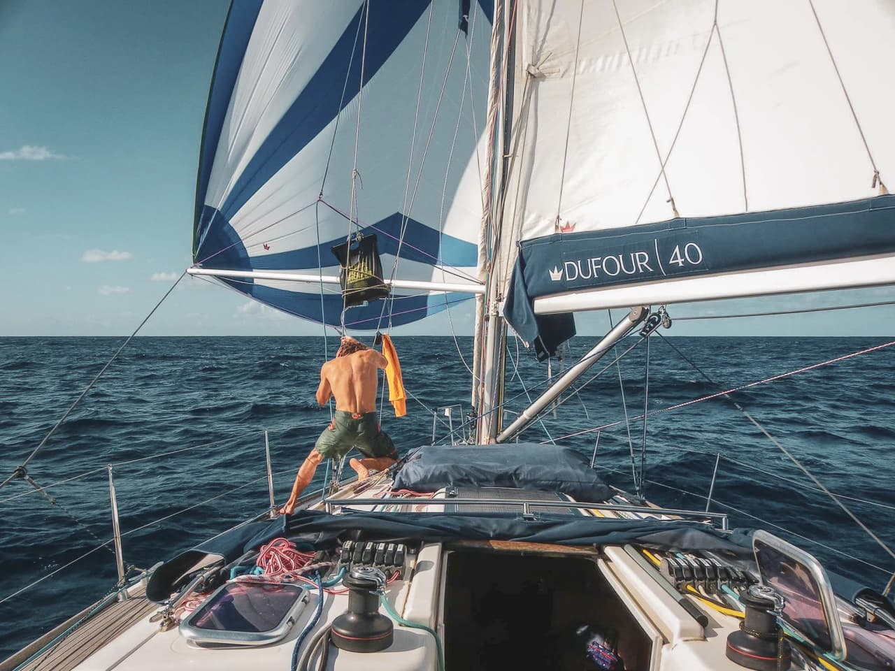 Un marin sur un voilier Dufour navigue en mer Méditerranée, avec des voiles colorées sous le soleil.