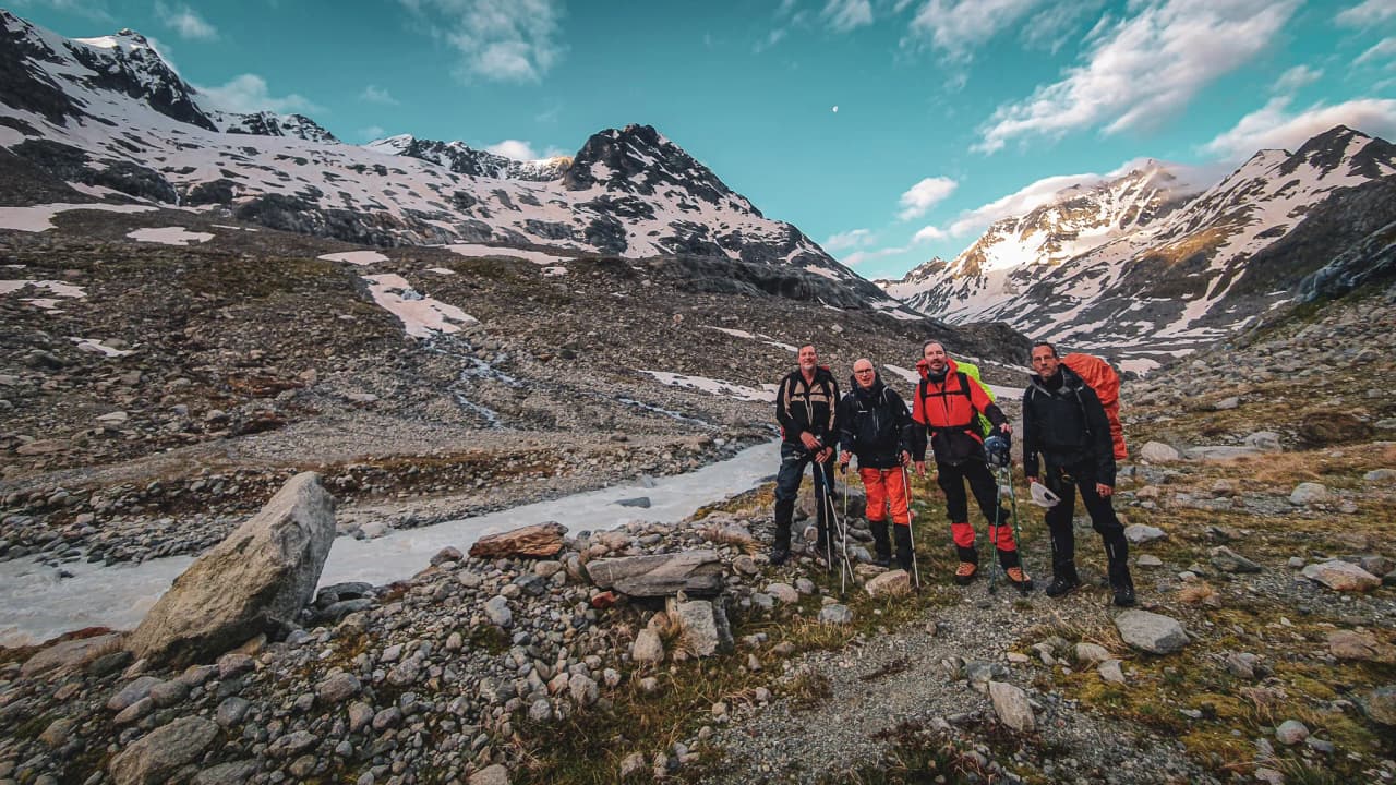 Four smiling hikers pose in a spectacular Alpine landscape, with mountains and glaciers in the background.