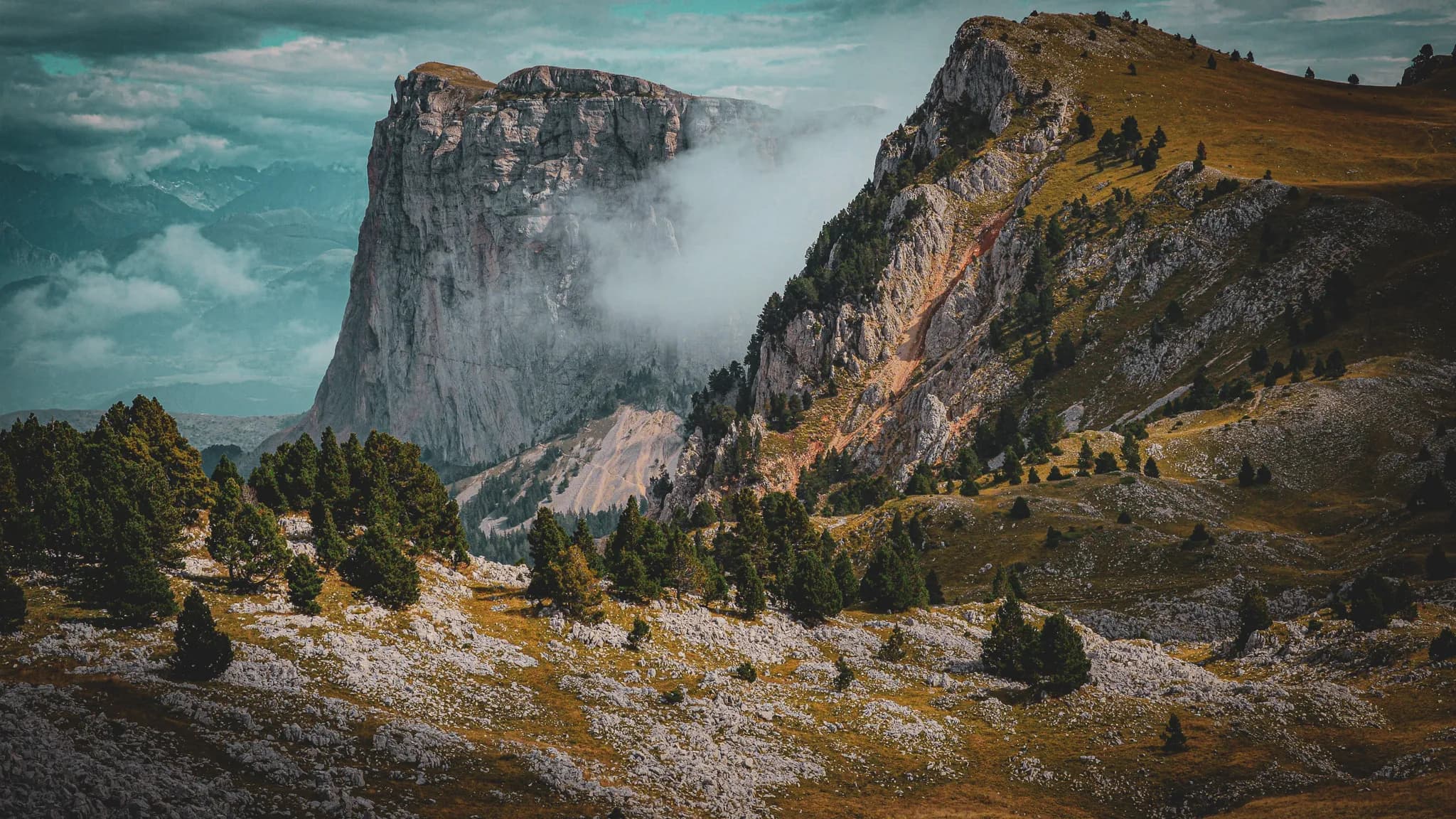 A steep mountain range rises majestically under a cloudy sky, with floating cumulus clouds. In the foreground, lush conifers cover a hilly, rocky terrain, while