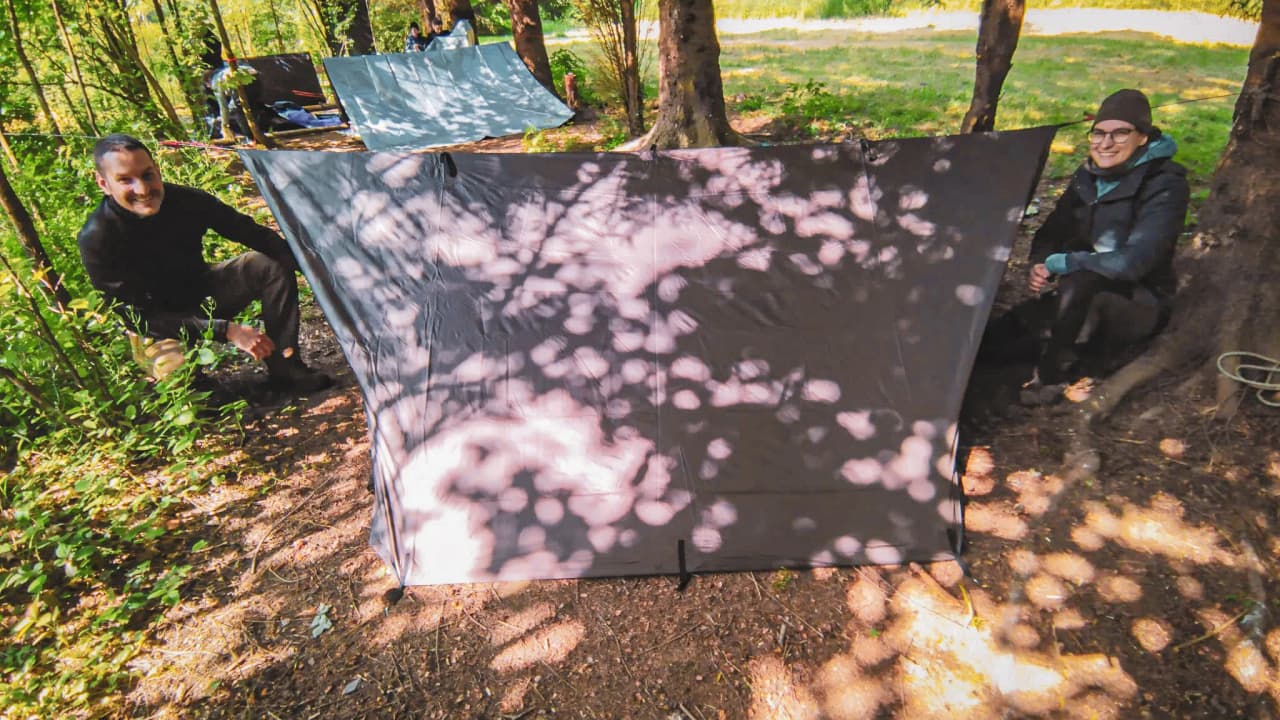 Deux personnes assises près d'un abri en forêt, entourées de verdure, prêtent de la survie en nature.