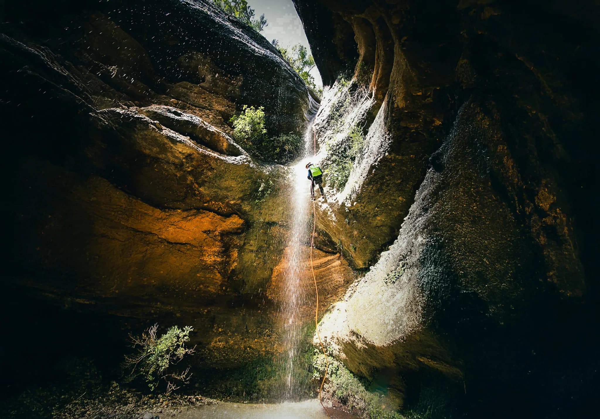 An adventurer descending rock faces in a canyon, with water flowing nearby. The walls are steep and surrounded by lush vegetation, while the ground is slightly