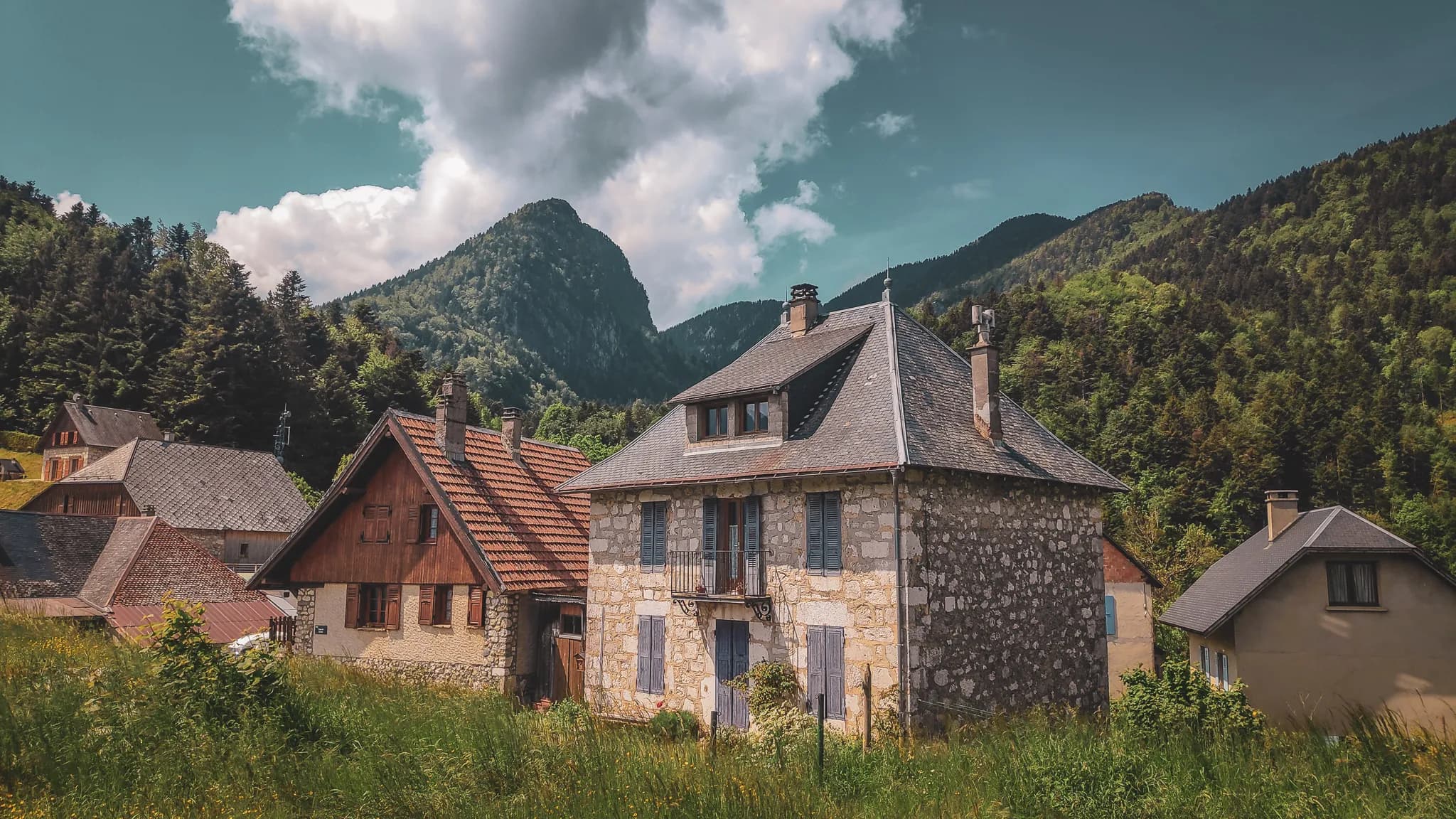 Un village pittoresque se niche au pied de montagnes verdoyantes, avec des maisons traditionnelles en pierre et en bois. La demeure centrale, dotée d'un balcon et de volets bleu, se distingue par sa façade claire, tandis que