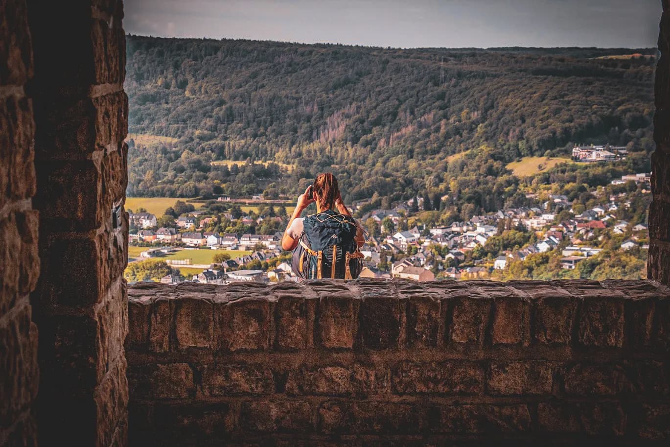 A person stands on a stone wall, gazing out over a green, undulating landscape. In the distance, houses are scattered across the valley, bordered by wooded hills. The light is soft, creating a