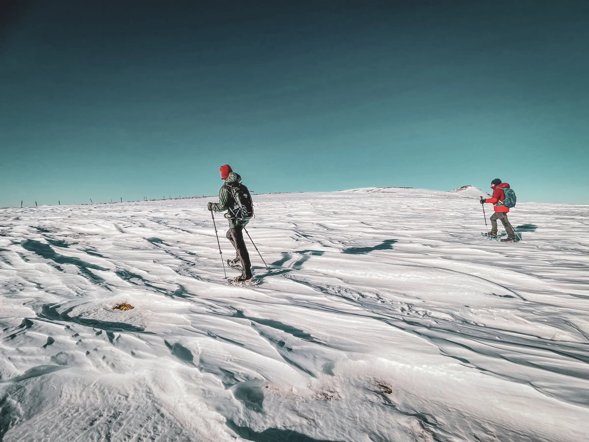 Twee mensen op sneeuwschoenen lopen door een besneeuwd landschap met rimpelingen in de sneeuw. De ene droeg een rode muts, een zwarte jas met groene accenten en een rugzak, terwijl de andere, op de achtergrond