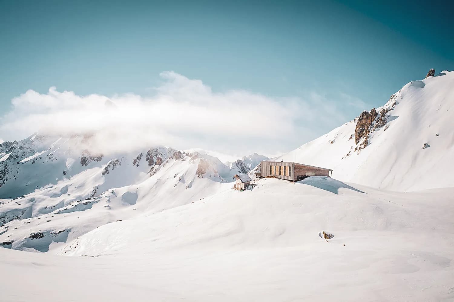 A modern wooden chalet stands on a snow-capped peak, surrounded by majestic mountains. The landscape is covered in a thick layer of snow, and hazy clouds float delicately over the