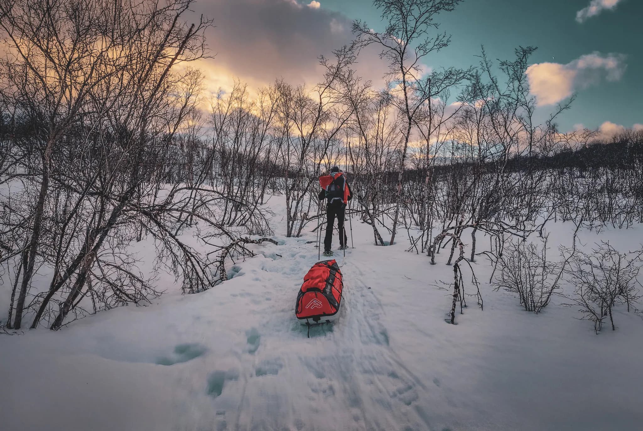 An adventurer in winter gear pulling a red pulka along a snowy trail, surrounded by bare trees and a wintry landscape. The sky is shades of orange and blue.