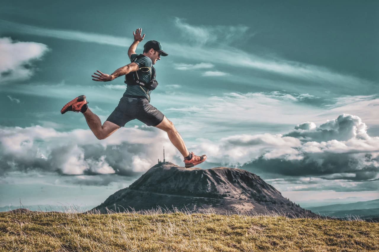 Un coureur de trail en plein saut devant un volcan majestueux sous un ciel nuageux.