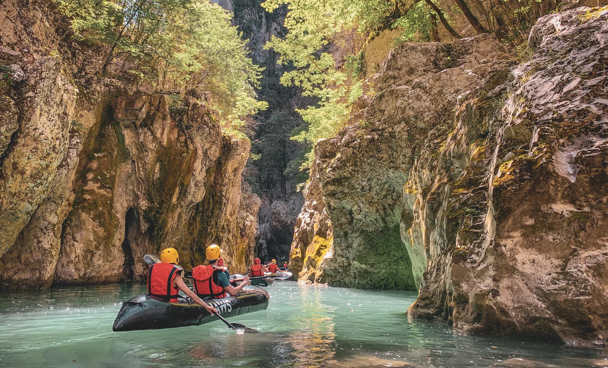 A narrow canyon lined with majestic rocks and lush greenery. Adventurers in kayaks calmly navigate the turquoise water, wearing orange life jackets and yellow helmets.