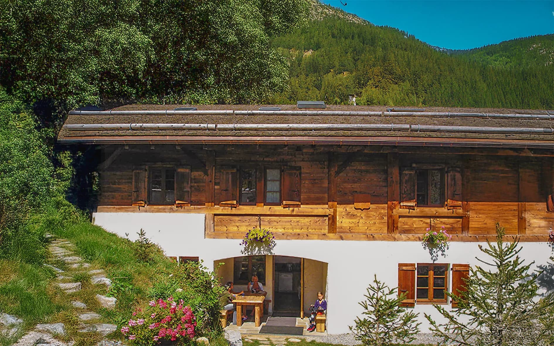 A wooden chalet is set in lush green countryside, surrounded by trees and mountains. The wooden façade is accentuated by wooden balconies and shuttered windows. On the