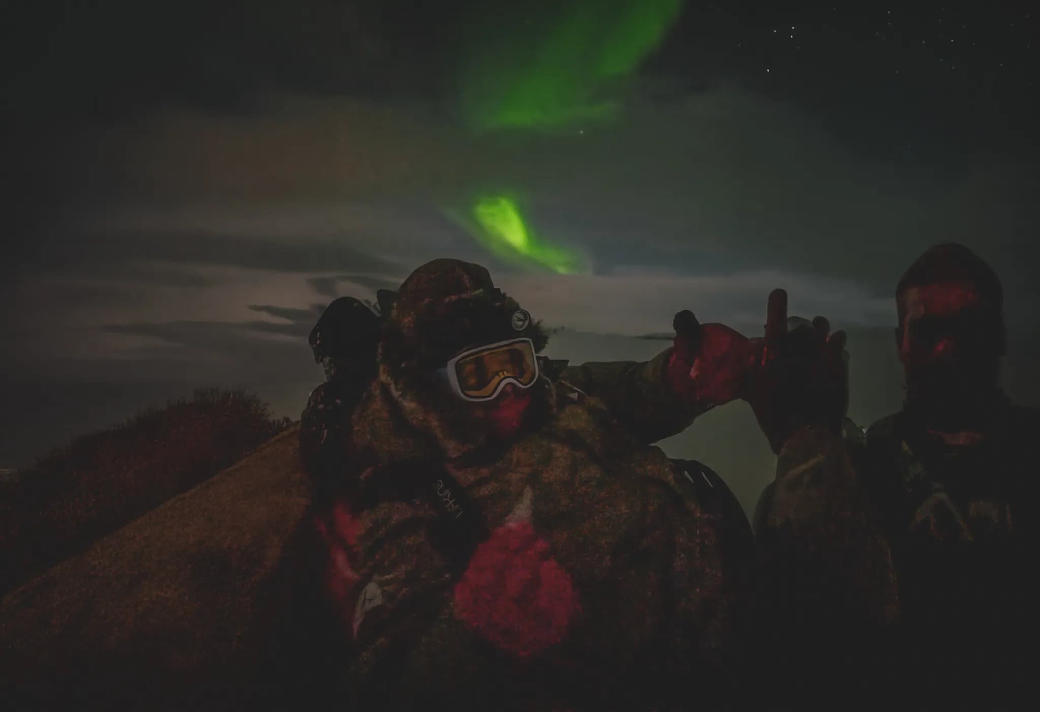A night scene with soldiers dressed in camouflage, resting on mountainous terrain. One of them is wearing goggles and appears to be waving. In the background, auroras
