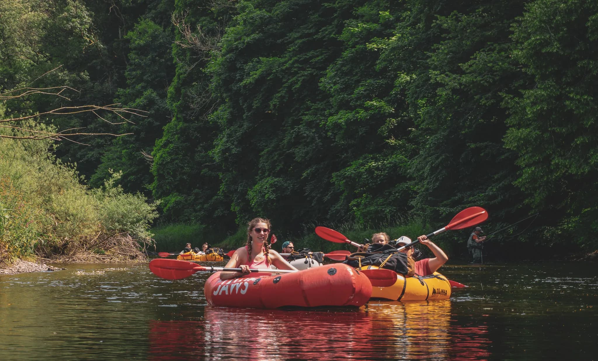 Twee kajaks drijven op een rustige rivier, omgeven door weelderig groen. Een van de kajaks is rood en draagt de naam "JAWS", terwijl de andere geel is met passagiers aan boord. Een vrouw