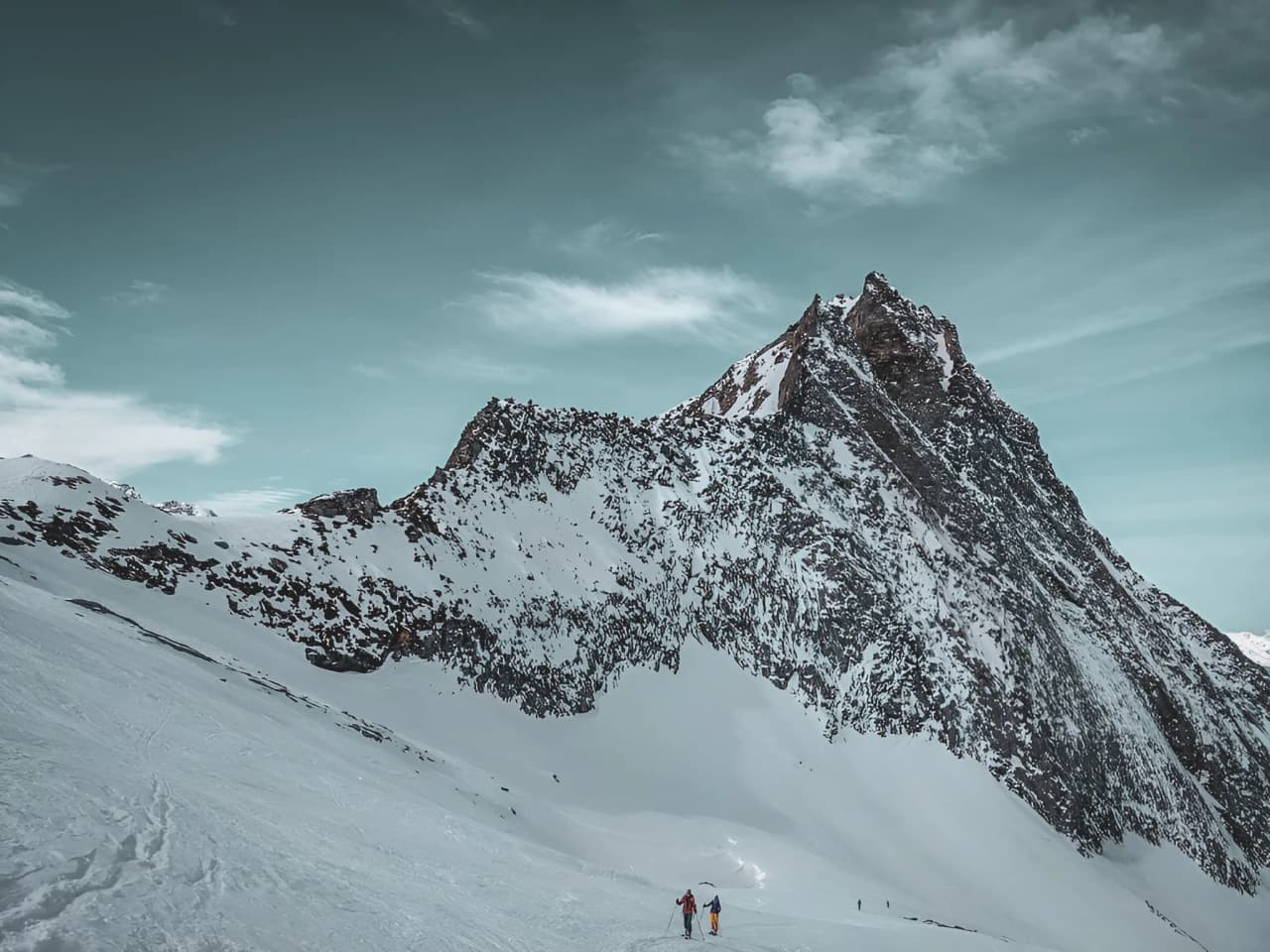 Two skiers make their way across a snow-covered alpine landscape, with a majestic peak in the background.