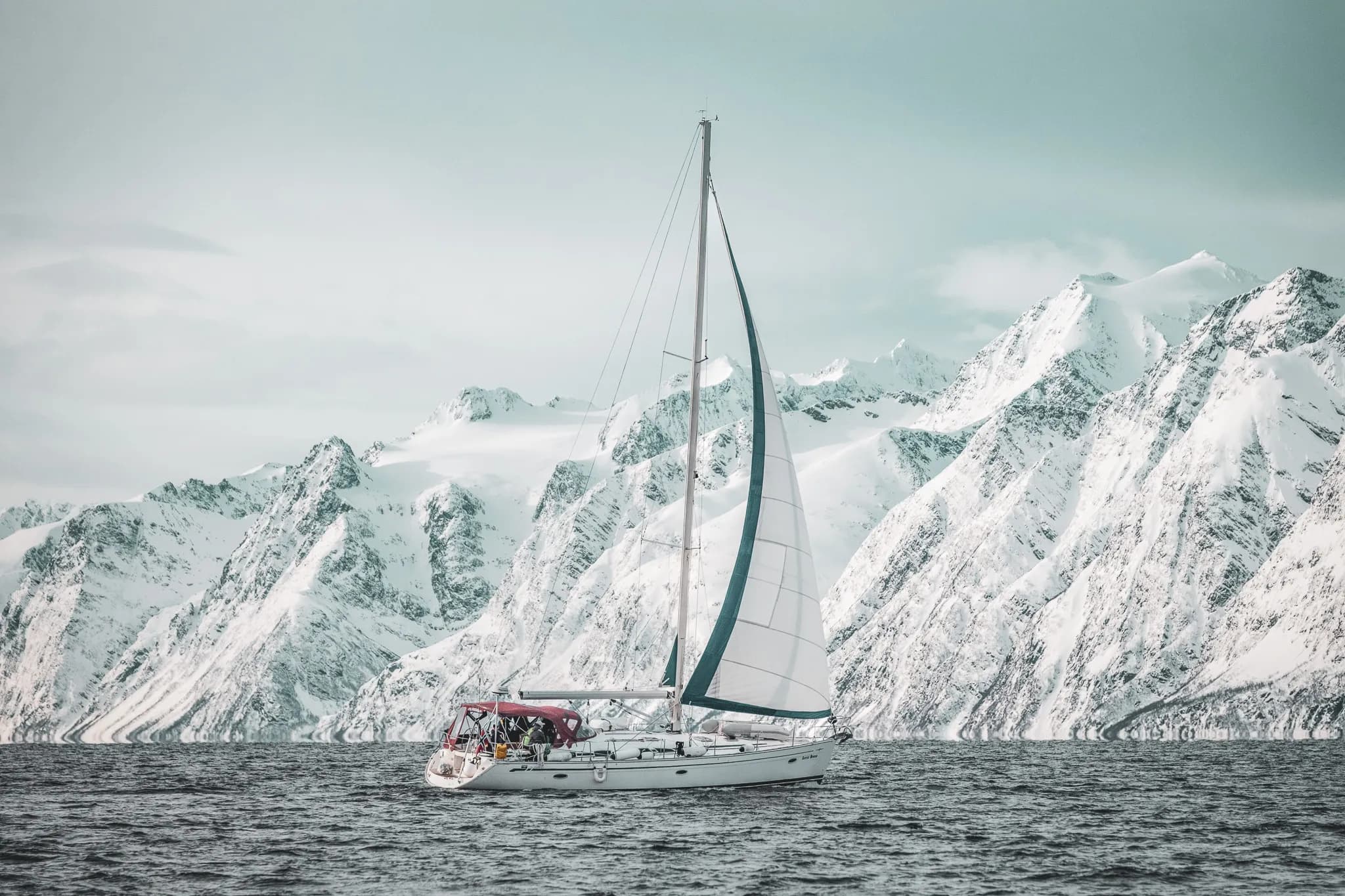 Un voilier navigue tranquillement sur des eaux calmes, entouré par des montagnes majestueuses recouvertes de neige scintillante. Le ciel est légèrement nuageux, offrant une lumière douce qui accentue les détails des sommets