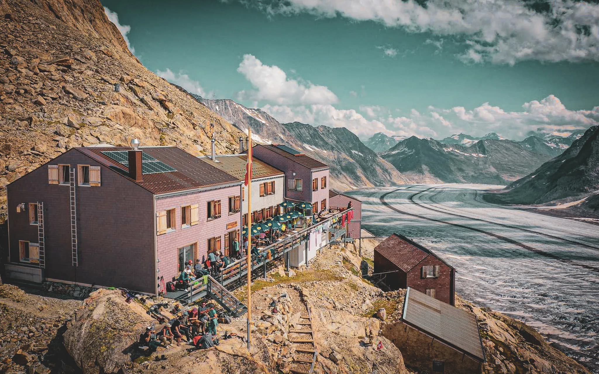 A mountain chalet built high above a vast glacier. The main structure is spacious, with large windows and a balcony where seats are arranged, welcoming people who seem to be enjoying the view.