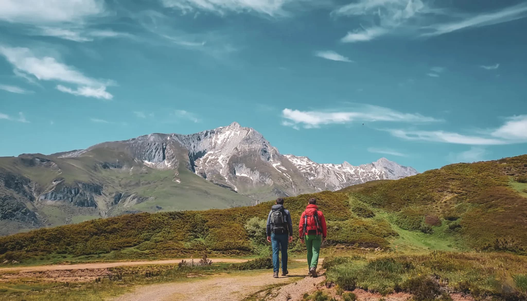 Deux randonneurs marchent vers les montagnes pyrénéennes sous un ciel bleu éclatant.