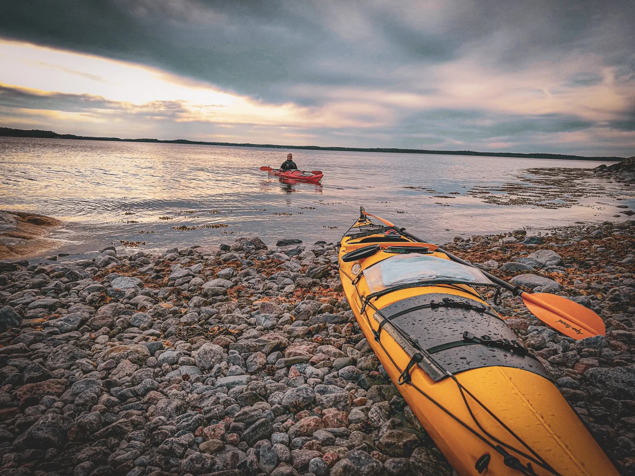 A yellow kayak rests on a rocky beach, while a second red kayak can be seen on the water in the distance, with a person sitting inside. The sky is cloudy and the sunlight is coming through softly at dawn.