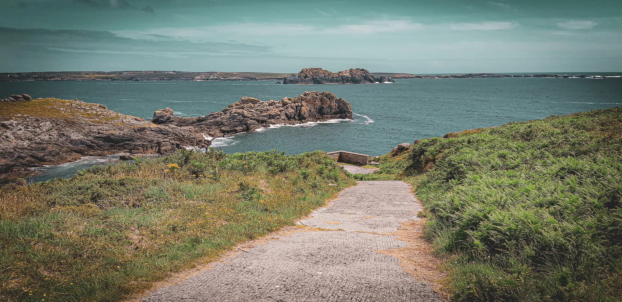 An engraved path leads gently down to the sea, surrounded by lush vegetation. Rocks emerge from the blue-green water, while the horizon is lined with green hills. The sky is slightly bare