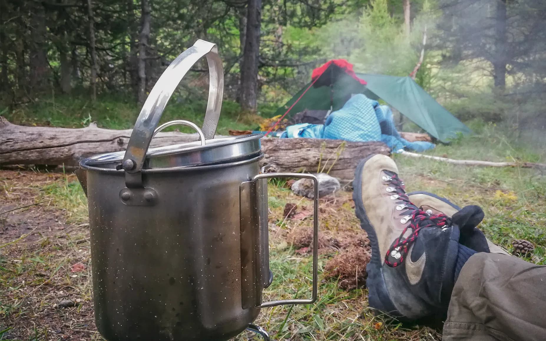 A metal pot smokes in the foreground, while a peaceful camp can be seen in the background.