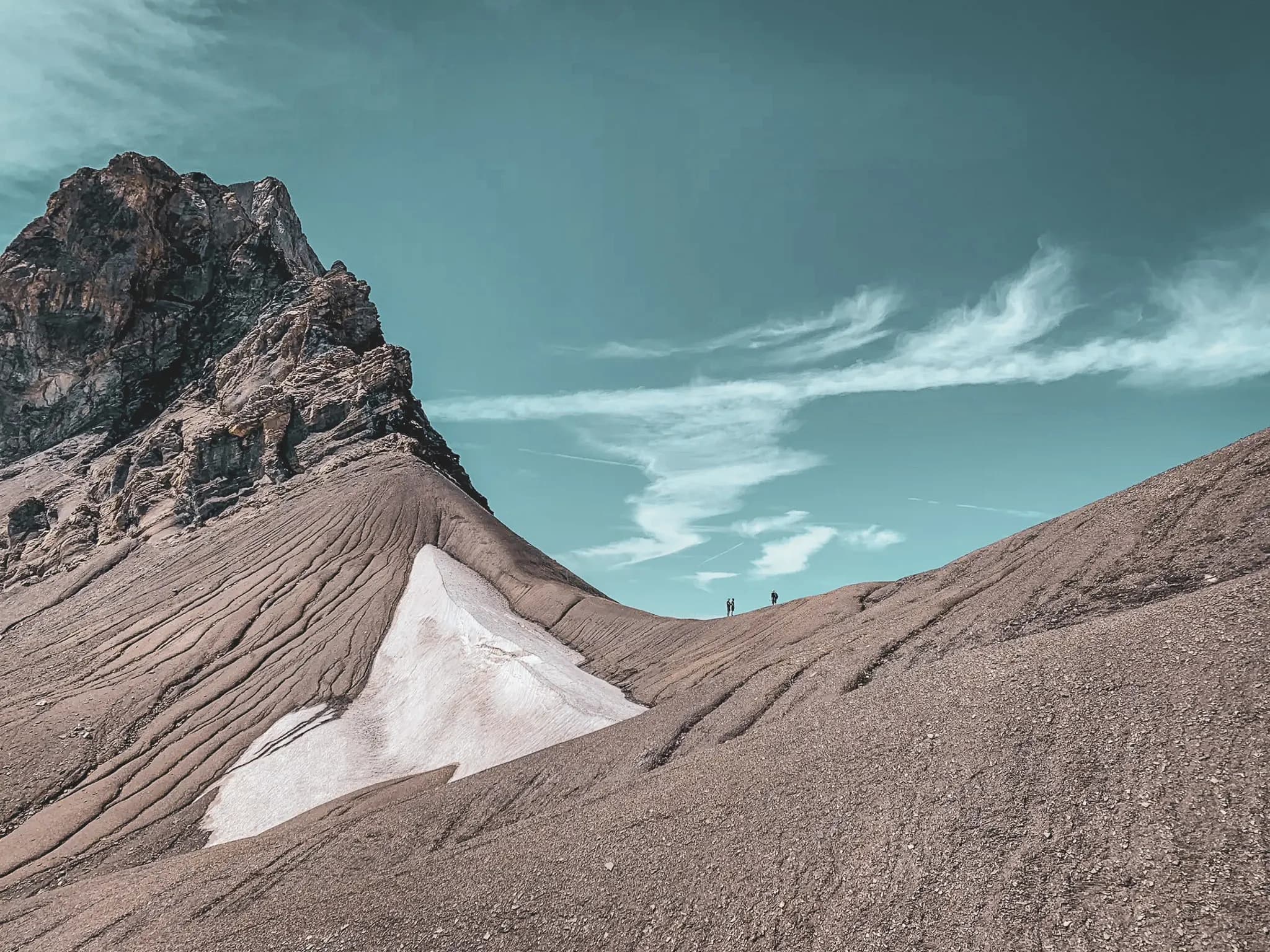A captivating mountain landscape with a steep, textured summit dominating a gentle slope. Streaks of snow stretch across the ground, contrasting with the brown hues of the earth. The sky is d