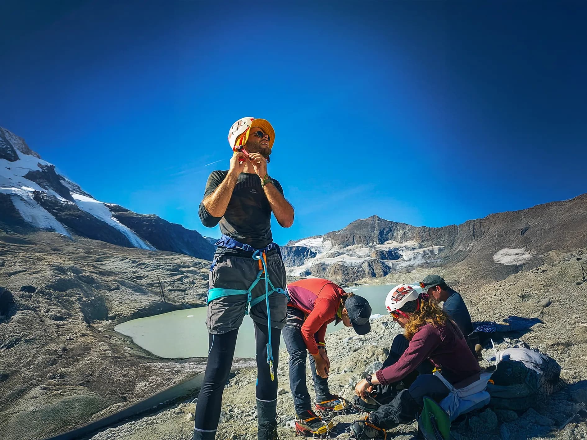 A group of people preparing for an outdoor activity in the mountains, under a clear blue sky. One of them is standing, adjusting his helmet and wearing a harness, while the others are looking after their equipment.