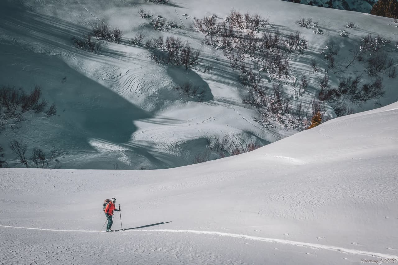 A lone skier moves through a snowy landscape, with a blanket of immaculate snow covering undulating slopes. In the background, shadows appear on the snow, creating a play of colours.