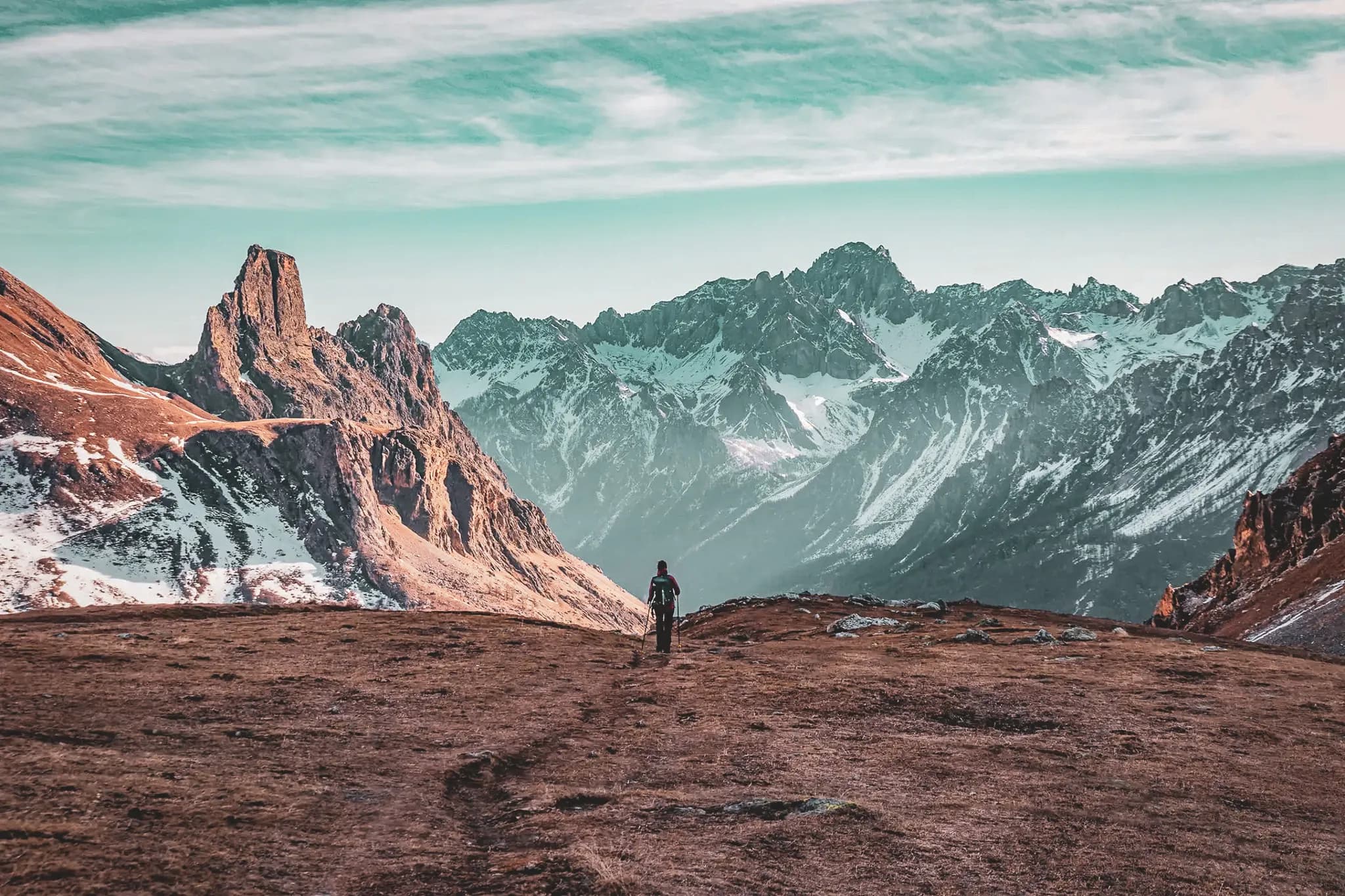 An impressive mountain landscape with snow-capped peaks in the background and a sky tinged with turquoise hues. A hiker, carrying a rucksack, heads towards the mountains, on a path winding through a