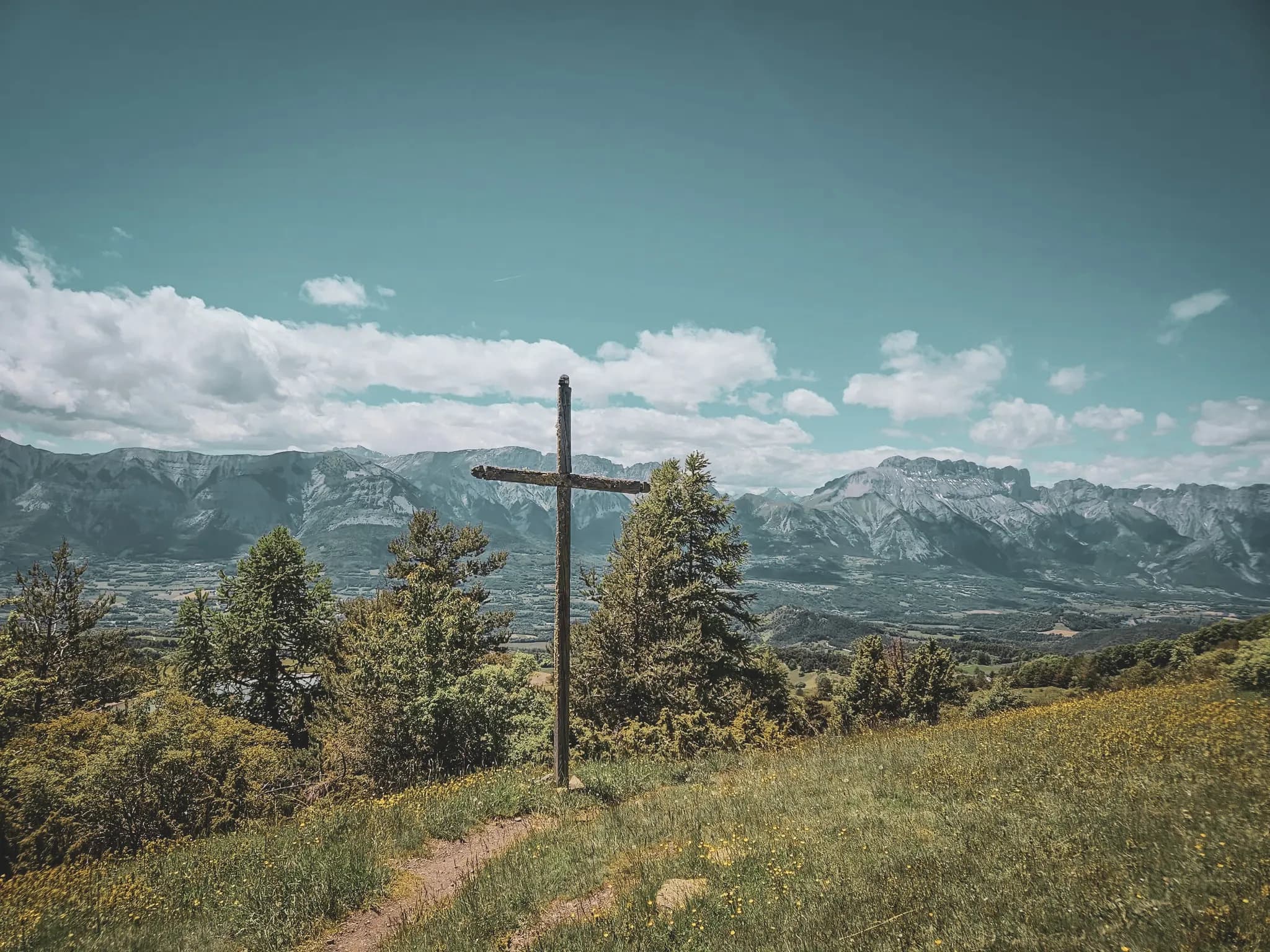 A tree-lined path leads to a wooden cross, with majestic mountains in the background and a serene sky.