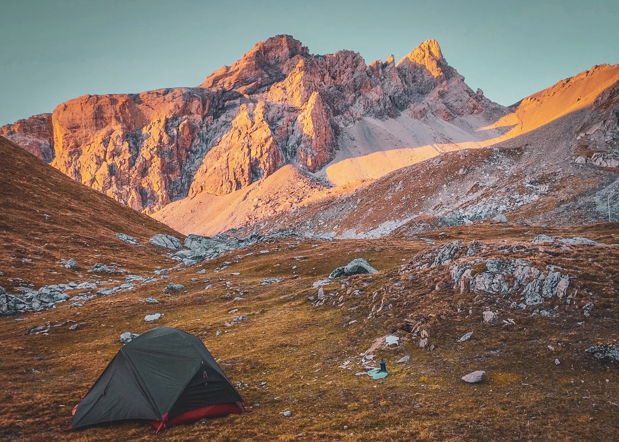 A campsite at the base of a majestic mountain range, with clear skies and warm hues lighting up the peaks. A dark-coloured tent is set up on a grassy, stony ground, surrounded by