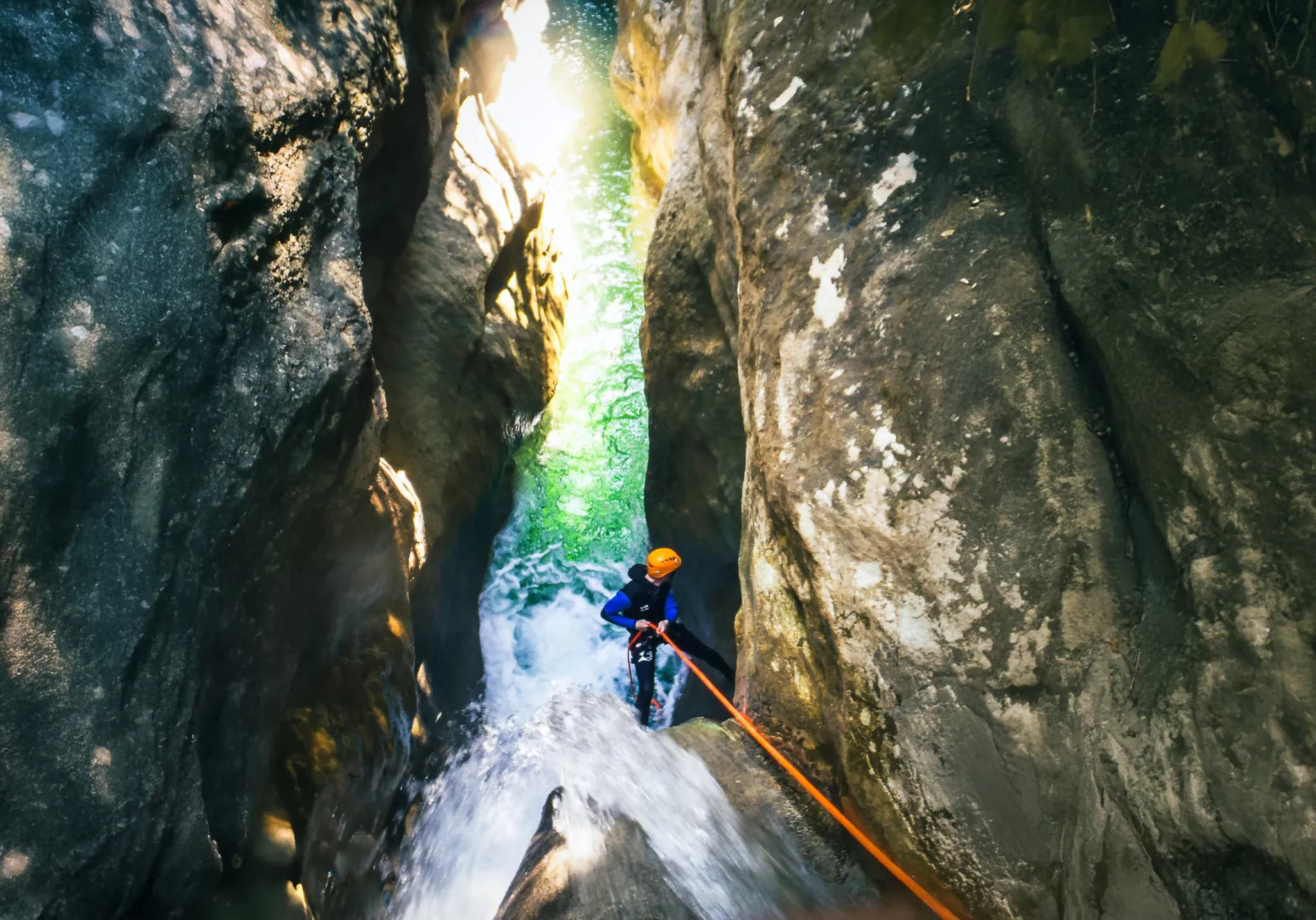 A man in a wetsuit and orange helmet descends a narrow rock face by rope, while a cascade of water gushes down at his feet. Sunlight filters through the cliffs