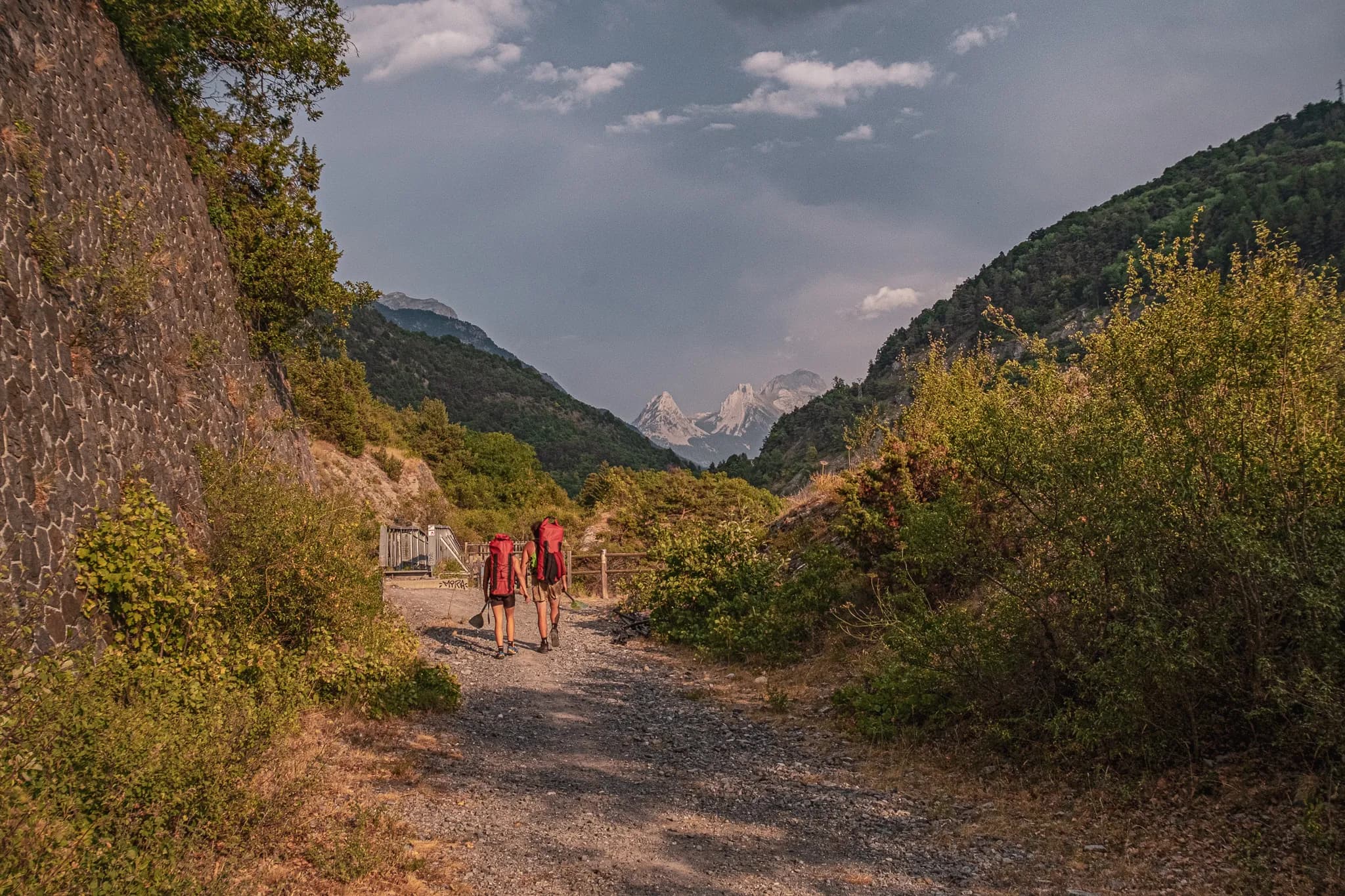 A gravel path winding between green hills, with two hikers carrying large red rucksacks walking side by side. In the background, majestic mountains rise up, their peaks covered in snow.