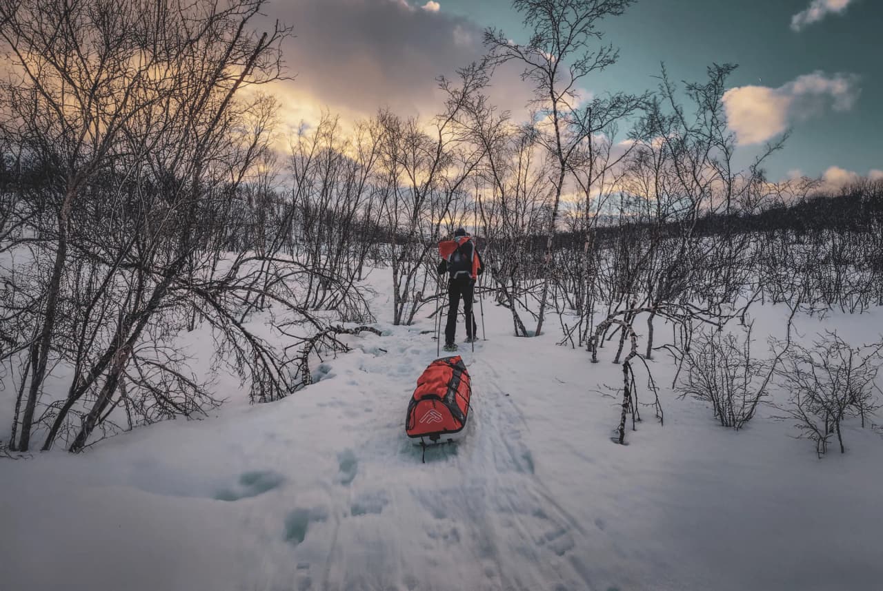 Snowshoe hiker on the Kungsleden, pulling a pulka, snow-covered landscape and colourful sky.