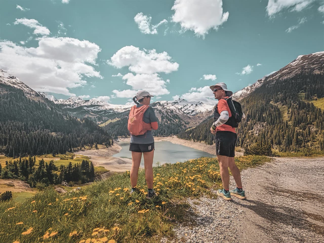 Deux randonneurs admirent un lac alpin, entourés de montagnes et de fleurs, sous un ciel lumineux.