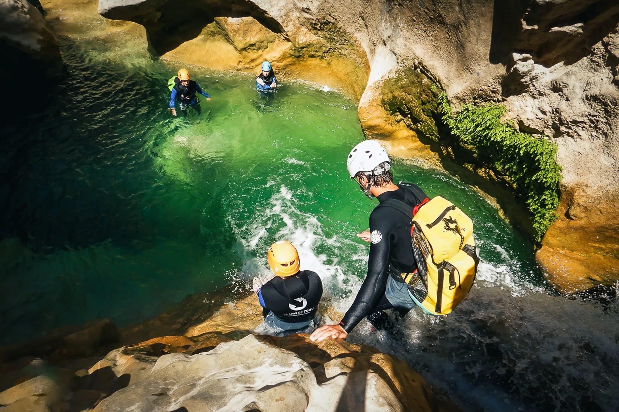 A group of people dressed in canyoning gear venture into a natural environment, with golden rocks and clear water tinged with green. One of the members, wearing an orange helmet, looks ready to go in.