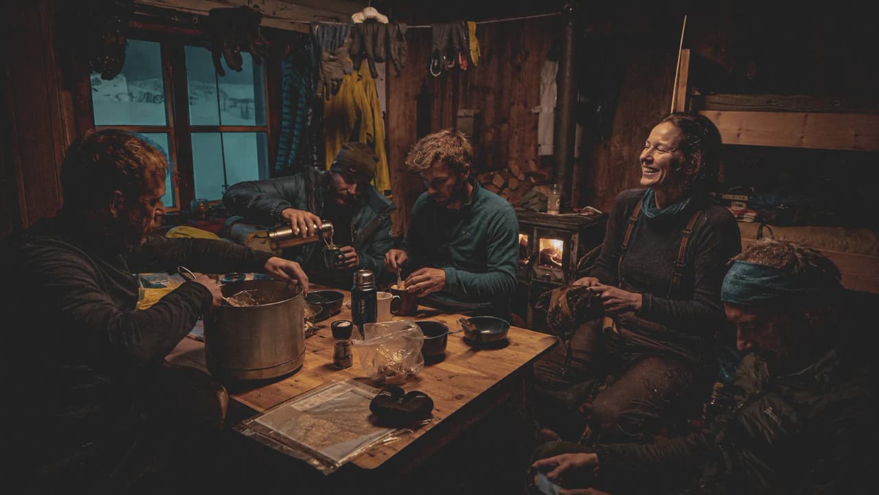 Vijf avonturiers genieten samen van een gezellige maaltijd in een met sneeuw bedekte Berghut, midden in de ongerepte natuur.
