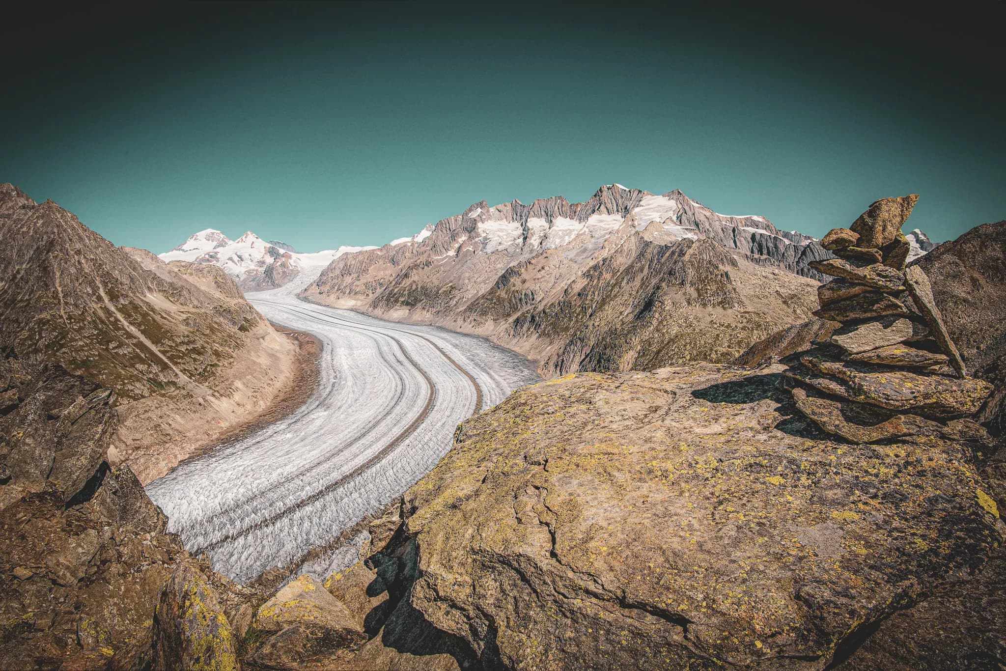 An impressive Alpine landscape with the Aletsch glacier winding its way through the mountains, surrounded by snow-capped peaks. Steep rocks, covered in moss and lichen, dominate the scene.