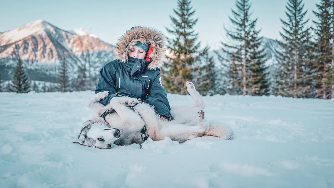 Een kind knuffelt een hond in een besneeuwd landschap, met bergen op de achtergrond.