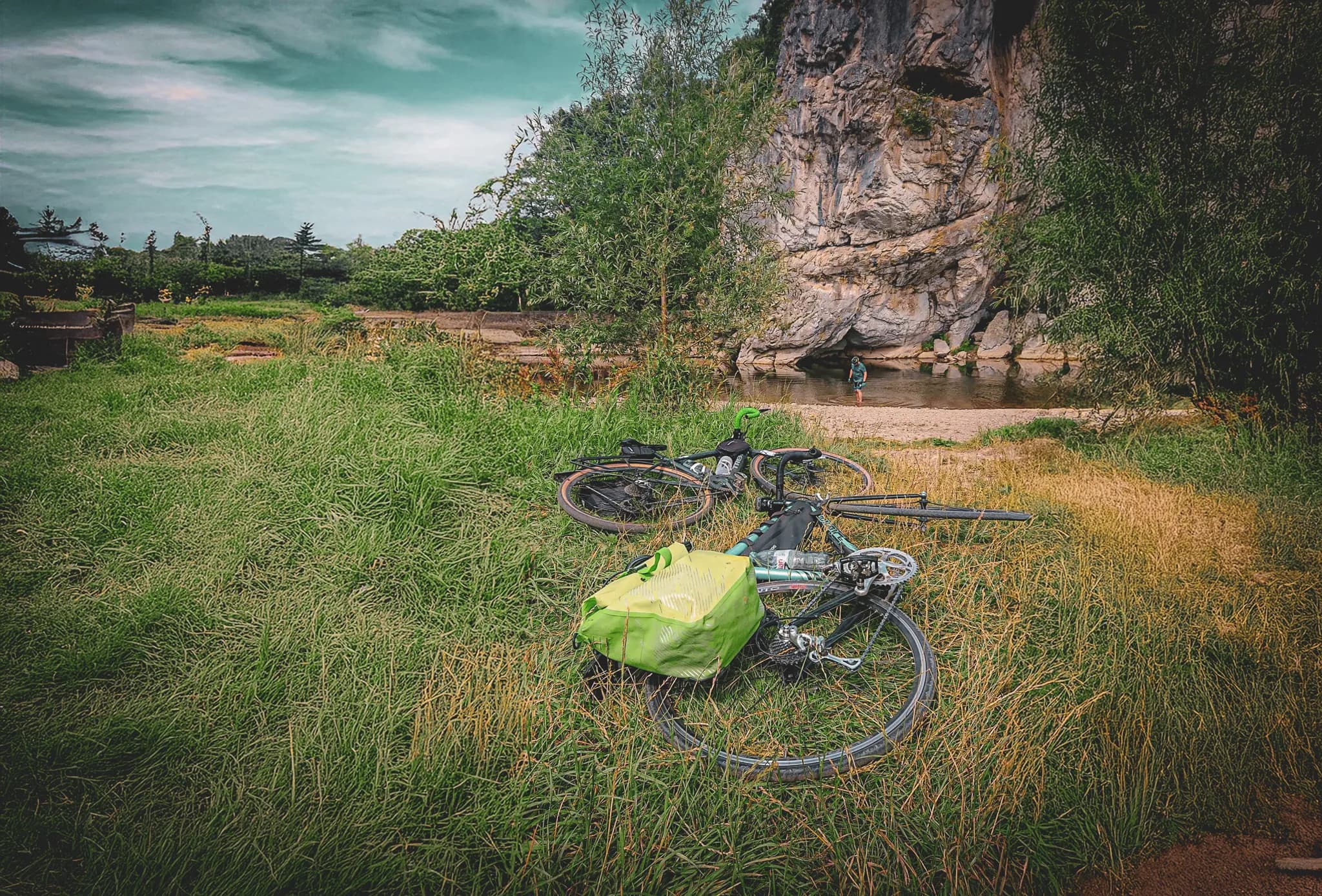 A natural landscape with dense green grass, where two bicycles lie on the ground. Nearby, a person stands in the shallow water of a river, enjoying the tranquillity of the surroundings. Visit