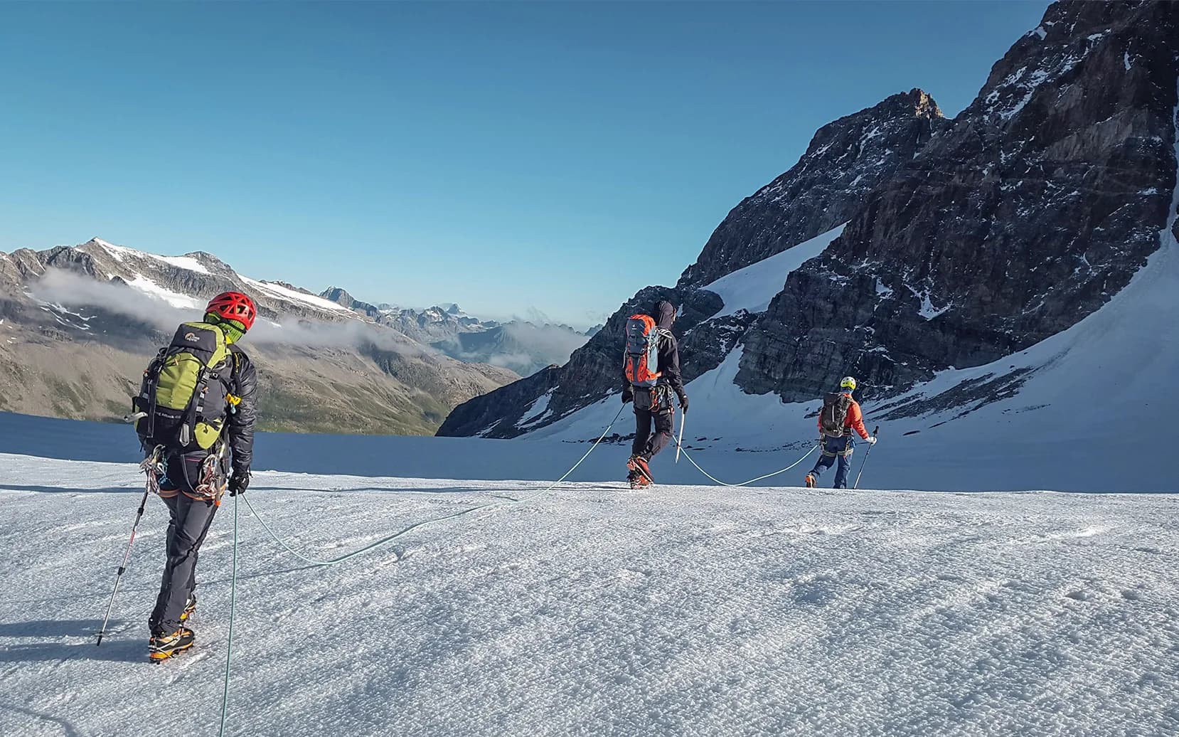 A group of three climbers on a glacier. They are equipped with mountain-climbing gear, including helmets and crampons. The surrounding landscape is spectacular, with mountain peaks