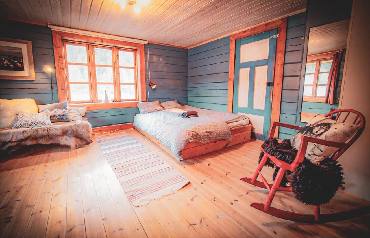 A cosy room with wooden walls painted in shades of blue and beige. A double bed, covered with a patterned duvet, is in the centre, with two pillows on top. Next to it is a small