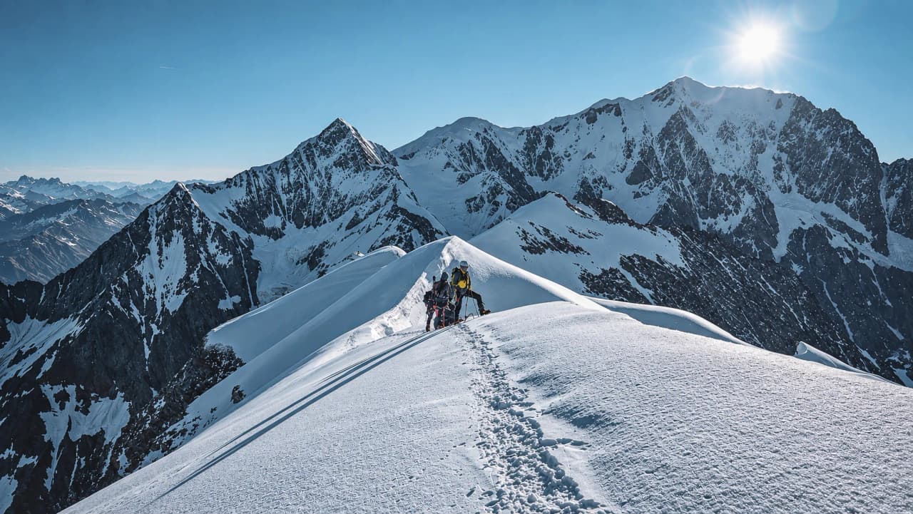 Drie bergbeklimmers klimmen naar een besneeuwde top met een spectaculair uitzicht op de bergen.