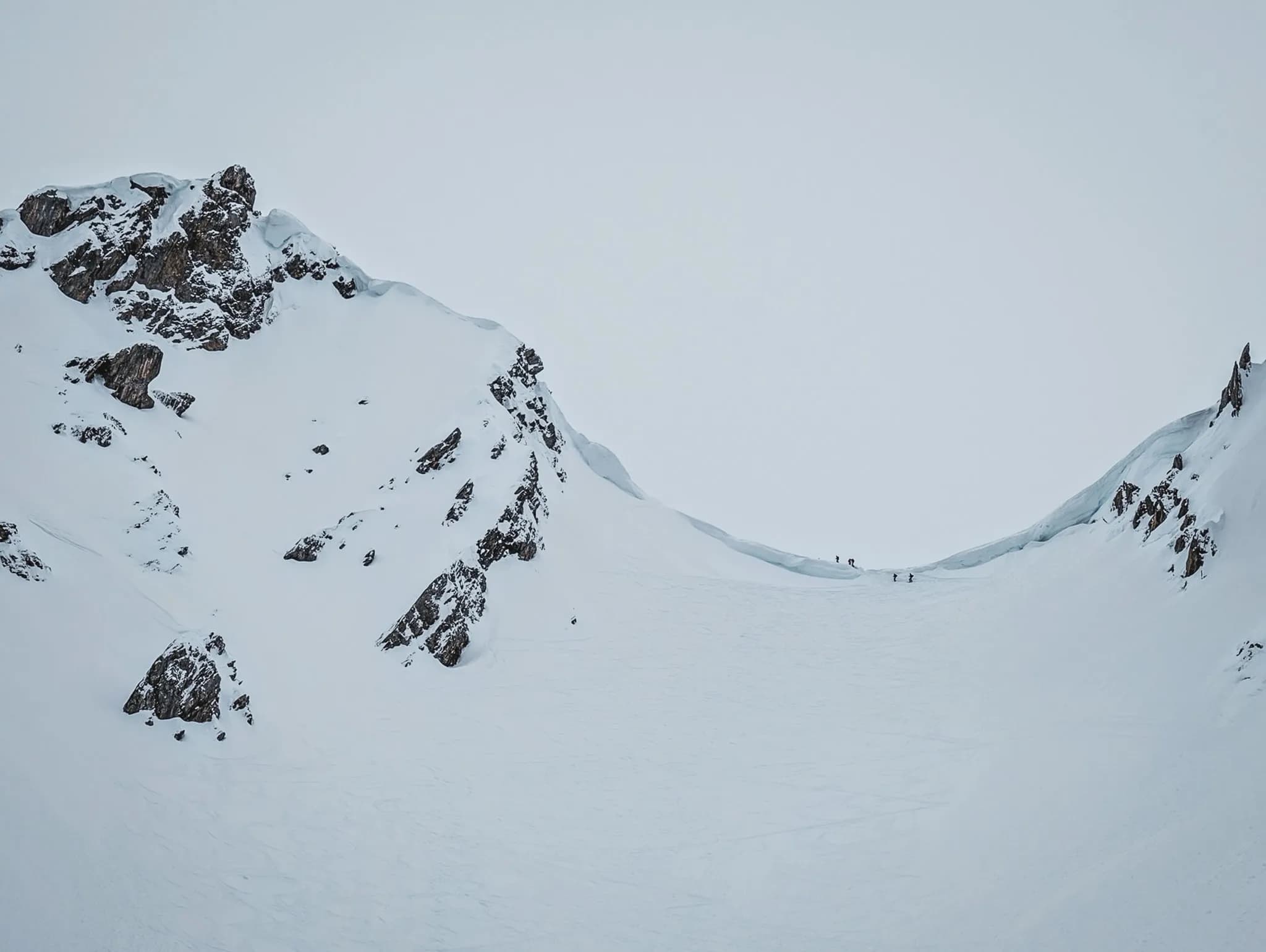 Un paysage montagneux enneigé avec des sommets rocheux en arrière-plan. La scène est dominée par un vaste champ de neige, parsemé de quelques rochers qui émergent. Au loin, on aperçoit de petites silhouettes humaines