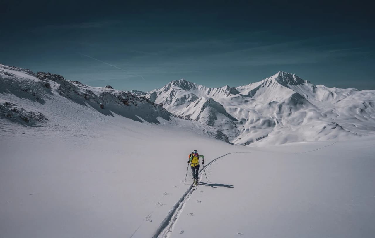 A lone skier crosses a vast snow-covered landscape, leaving tracks in the immaculate snow behind him. Majestic mountains rise up in the distance, their peaks covered in snow. The sky is blue